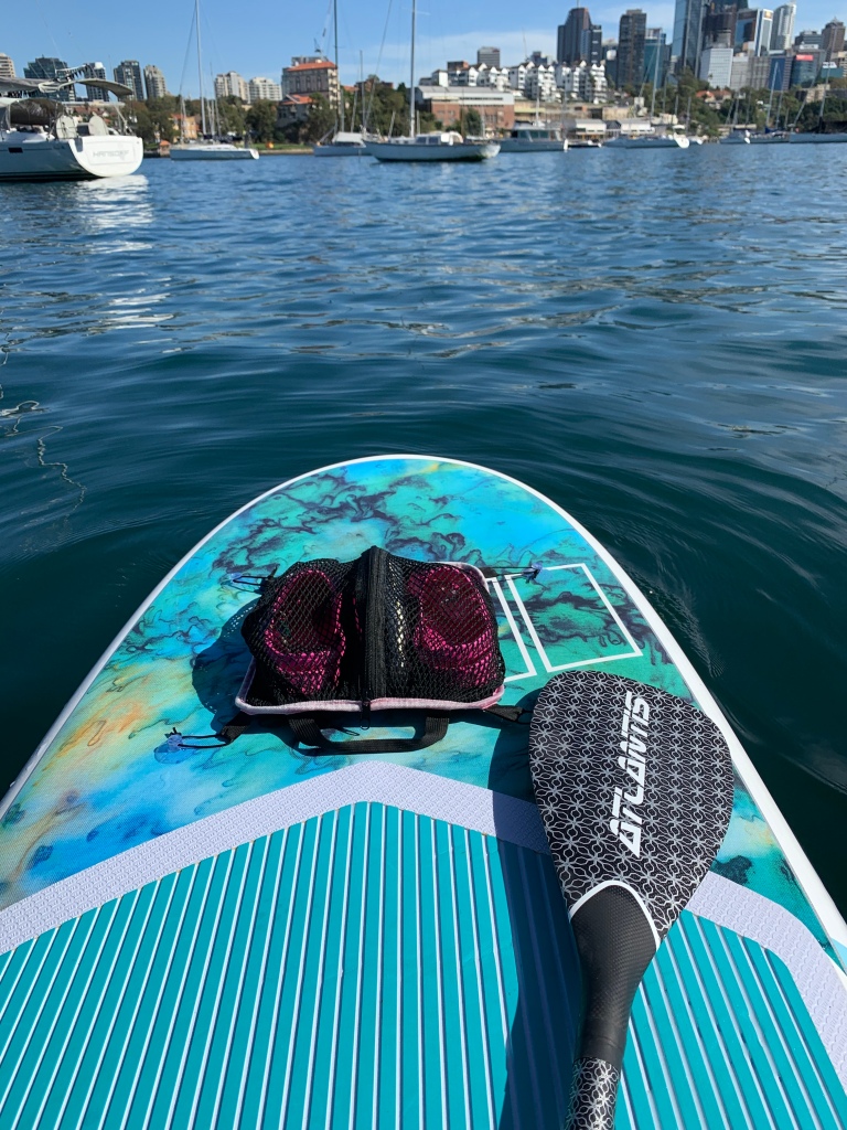 A pair of pink crocs are secured to the front of a stand up paddleboard floating in calm waters